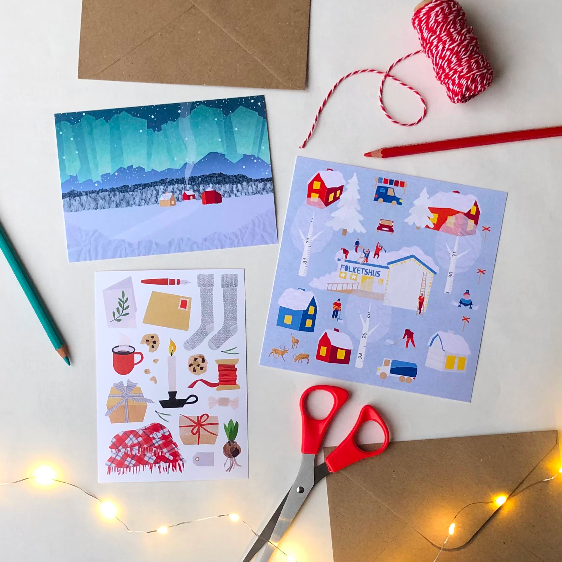 A flatlay view of 3 postcards with envelopes, styled with a bobin of red and white twine, colored pencils, a pair of cissors and a string of lights.